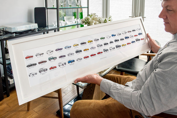 Person holding a framed artwork with various car illustrations in a room.