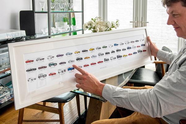 Person holding a framed print of various cars in a room with a glass cabinet.