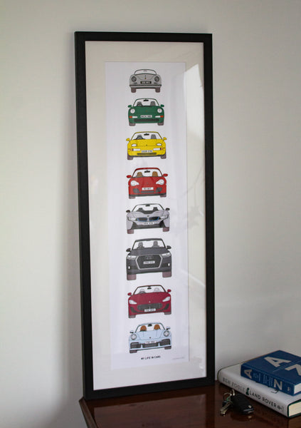 A tall black-framed art print featuring a vertical sequence of classic car illustrations in different colors, leaning against a light wall on a wooden surface next to books.